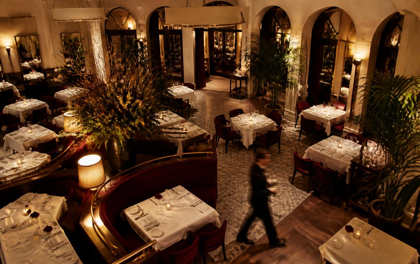 An elevated view of a dining room in service, a server moving through the room between set tables.